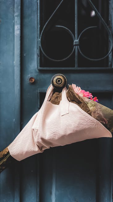 A person wearing a latex glove holds a bouquet of fresh pink roses wrapped in light pink paper with a textured surface and tied with a brown paper wrap. The bouquet is being delivered to a dark blue d