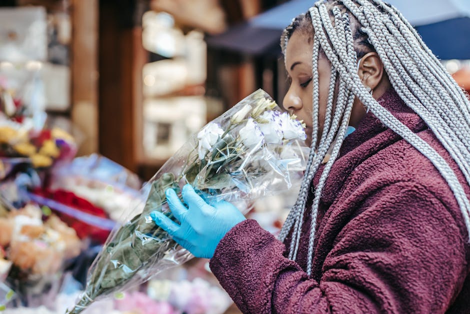 A woman with long, braided silver hair wearing a maroon fleece jacket and blue gloves holds a large bouquet of white roses wrapped in clear plastic. She appears to be smelling the flowers at an outdoo