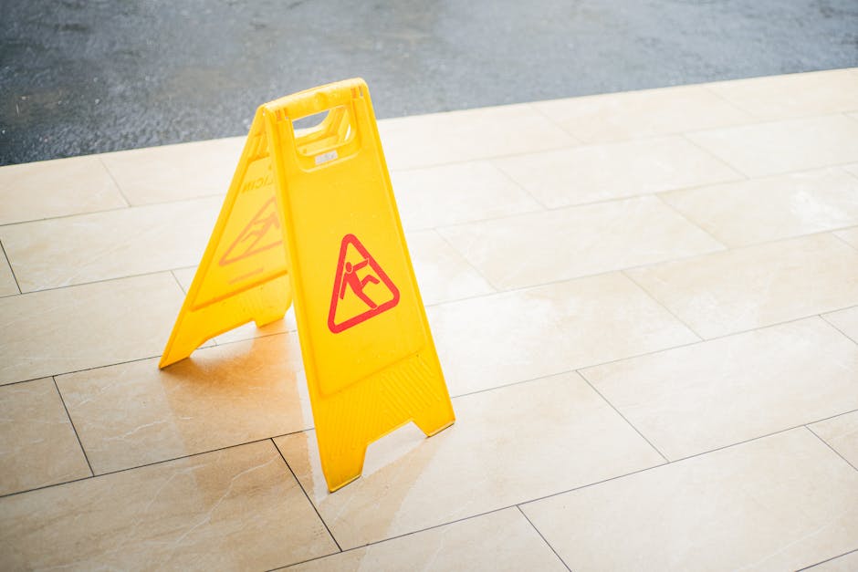 A bright yellow, portable floor sign with red warning symbols indicating a wet floor, placed on beige tiled flooring near an entrance or corridor. The sign stands upright and is illuminated by natural