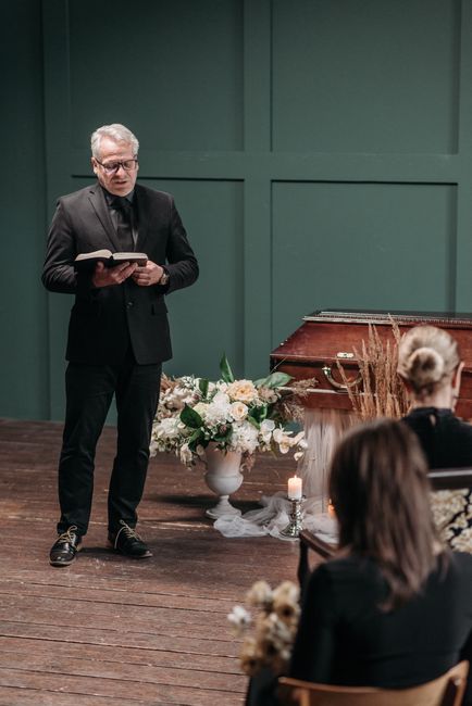 A man with grey hair and glasses, dressed in a black suit, is reading from a book during a funeral or memorial service held indoors. In front of him, there is a large white urn-style floral arrangemen
