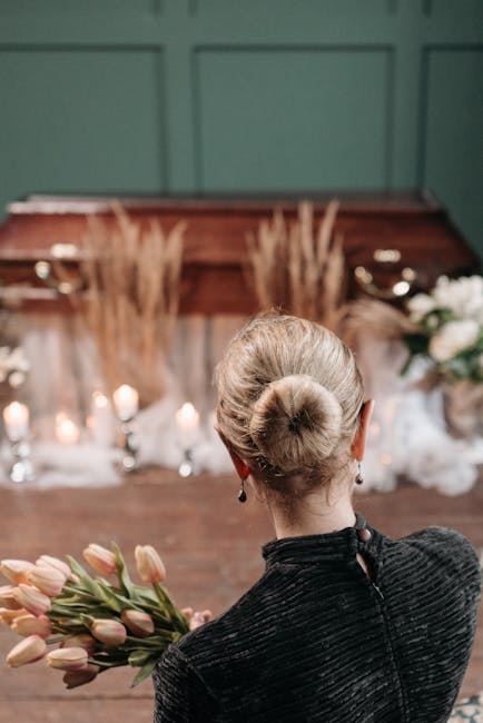 A woman with platinum blonde hair styled in a neat bun, dressed in a black textured jacket, holds a bouquet of pale pink tulips with green stems and leaves. She stands indoors, facing away from the ca