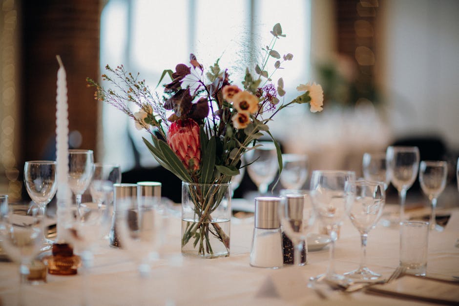 A floral arrangement featuring a mix of fresh flowers including protea, small daisy-like blooms, and filler greenery arranged in a clear glass vase positioned at the center of a set dining table. The 
