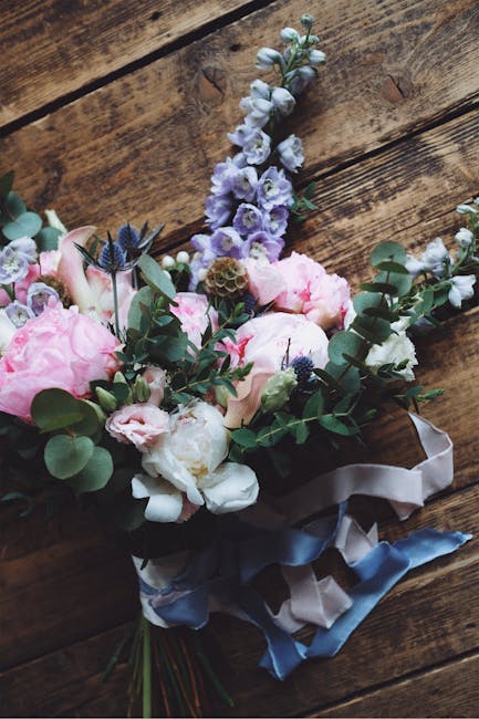 A floral bouquet arranged with large, soft pink peonies, white and pale purple delphiniums, and sprigs of eucalyptus with rounded green leaves. The flowers are freshly cut, exhibiting textured petals 