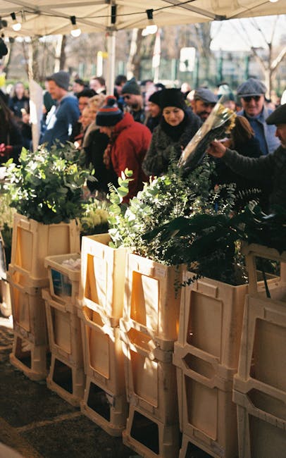 At an outdoor flower market under a beige canopy, several wooden crates filled with fresh greenery and floral arrangements are displayed. The flowers include lush green foliage, delicate eucalyptus le