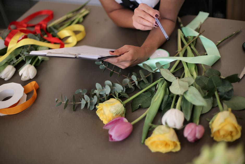 A person at a work table arranging a floral gift, surrounded by various fresh flowers including yellow roses, pink tulips, and white tulips, along with eucalyptus leaves and green stems. The person’