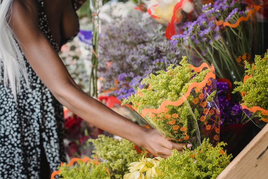 A person with dark skin and long blonde hair, dressed in a black and white polka dot top, arranging a bouquet of fresh flowers at a flower shop. The bouquet features vibrant green ornamental grasses, 