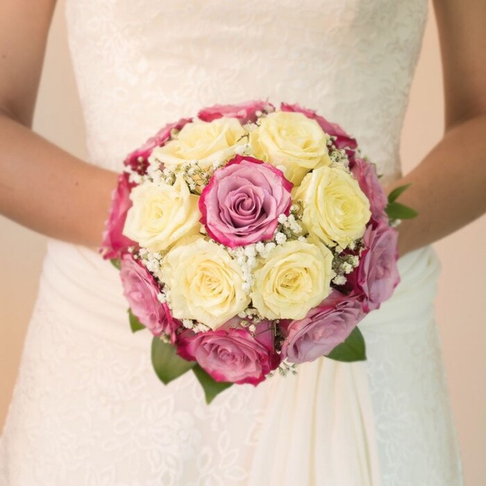 Immersed in soft, even lighting reminiscent of a South Hackney afternoon, this eye-level photograph draws close to a bride's torso, her arms encircling a superbly crafted round bouquet. The floral arrangement bursts with an array of creamy lemon-yellow roses, each bloom unfurling in perfect succession, complemented by vibrant fuchsia roses and blush-mauve shades that bring contrast and depth. Delicate sprays of pristine white baby's breath weave in and out between the roses, adding airiness and subtle texture to the overall composition. Hints of lush dark green foliage peek from beneath the blooms-an echo of the abundant greenery that lines Mare Street and nearby Hackney Marshes. The bride's understated elegance is further highlighted by the lace embellishments across her gown's bodice and the gentle drape of flowing fabric below, all gently illuminated by clear daylight. The bouquet radiates a mood of celebration and meticulous artistry, capturing the unique character of South Hackney weddings-where traditional English blooms meet contemporary style and local creativity. This image stands as a tribute to the expertise and pride of South Hackney's skilled florists, a touchstone for those seeking meaningful design on their most important day.