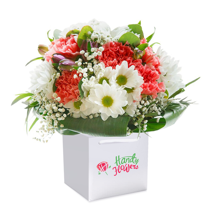 A vision of contemporary floristry, this vibrant bouquet commands attention from atop a spotless, modern white gift bag set against a pure white background-flawless and inviting. The arrangement itself is a harmony of lush coral and peach carnations, their abundant, frilled petals forming luxurious, textured clouds that breathe energy into the scene. Interspersed throughout are crisp white daisy-like blooms crowned with striking yellow-green centers, their cheerful presence reminiscent of Hackney's thriving gardens and community parks. Tiny stars of delicate baby's breath create a sense of lightness, artfully threaded among the larger blossoms for an airy effect. Toward the bouquet's left edge, a few unopened buds with purple-tinged hues add subtle dimension and the promise of more blooms. A collar of generously broad, deep green leaves surrounds the base, their lustrous surfaces framing the flowers with elegance-a nod to the leafy stretches along Hackney Downs. The arrangement, rounded and full, radiates freshness and meticulous craftsmanship, all thoughtfully contained within a gift bag designed for convenience-complete with robust white ribbon handles for comfortable carrying from Dalston to Homerton. Ideal for the varied celebrations and thoughtful gestures that mark life in South Hackney, this bouquet beautifully blends expert technique, natural abundance, and a sense of local pride. There's no mistaking the care and personality infused by South Hackney's finest florists in every stem and petal.