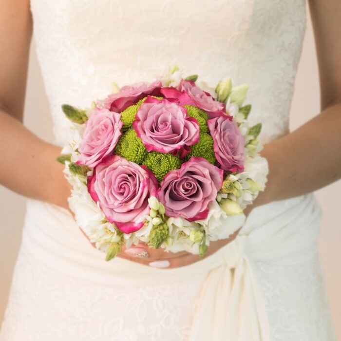 From a waist-level angle, this photograph immerses us in the South Hackney wedding experience: a bride, poised and serene, holds a vibrant bouquet in full view over her classic, off-white dress near the lively heart of the borough. The floral arrangement is a testament to local craftsmanship, its round shape drawing the eye to the plush roses at its core-petals fading from a soft lavender-pink into arresting fuchsia at the tips, each bloom inviting admiration. Between the roses, vivid lime-green button chrysanthemums-tightly packed and springy-inject the bouquet with a dash of unexpected exuberance reminiscent of the fresh grasses at Haggerston Park in early summer. Surrounding these central flowers is a border of delicate white blossoms, most likely clusters of freesia or tiny spray roses, their green buds and slender leaves completing an aura of freshness and natural grace. The subtle gleam from the bride's light pink polished nails with a hint of sparkle complements the bouquet's palette. Illuminated by gentle daylight and the soft ambience typical of Hackney venues, this bouquet brings together artistry, tradition, and the distinctly contemporary style of weddings in South Hackney. It's a floral tribute crafted for memorable moments in settings from Bethnal Green to London Fields.