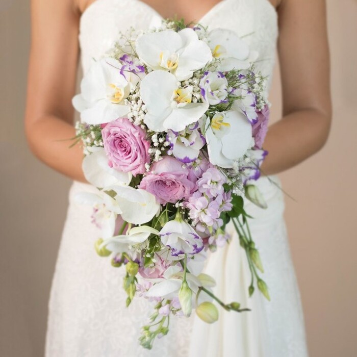A stunning, vertically composed close-up reveals a South Hackney bride gently holding a lavish, cascading bridal bouquet in front of a softly blurred beige backdrop. Shown from chest down, she wears an elegant white strapless gown, its bodice and skirt alive with detailed lacework, reminiscent of ceremonial finery often seen near St John's Church or Hackney Town Hall. Her bouquet unfurls in a graceful teardrop shape, the eye immediately caught by large, velvety phalaenopsis orchids-pristine white with glowing yellow and faint purple centers. Lush, blush-pink garden roses nestle in their midst, their petals softly curled, complemented by slender stalks of lavender stock and lisianthus, some edged in deeper violet. Wisps of baby's breath create an airy softness, while careful touches of greenery and trailing stems add organic movement. The bouquet's dramatic yet refined silhouette, paired with the bride's gentle posture, radiates the artistry of South Hackney florists. Whether destined for a stylish wedding at the Geffrye Museum or an intimate bash along Well Street, this bouquet combines local charm, sophistication, and floral storytelling in a single, breathtaking vision.