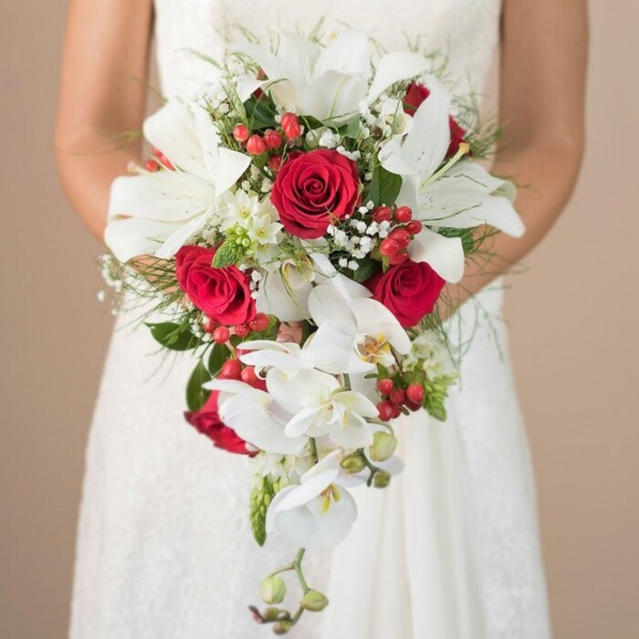 In this richly textured image, a South Hackney bride-her white lace wedding gown partially visible-holds a breathtaking shower-style bouquet designed with local artistry in mind. Dominant are the pure white oriental lilies, their open, trumpeted forms exuding both strength and elegance, while trailing white phalaenopsis orchids provide a sense of natural movement, leading the eye downward in a graceful waterfall effect. Lush, sensuous red roses bloom among the white, their deep hues echoing the vibrance of Hackney's urban energy, and small glossy red hypericum berries punctuate the composition with brightness. Clouds of white gypsophila and petite filler blossoms soften the transitions, edged by wispy asparagus fern and glossy green leaves that speak to the abundant life of parks such as London Fields. The bride's bare arms are seen warmly embracing the bouquet, while the tailored lacework of her gown hints at Hackney's blend of old and new. This bouquet from South Hackney is an artful tribute to local creativity-a meaningful choice for modern couples marking their vows within the borough.