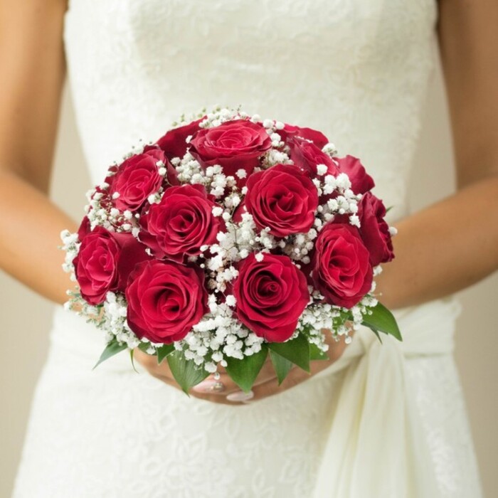 Lit by the gentle rush of day across South Hackney, this mid-torso close-up beautifully captures a bride presenting a domed, elegant bouquet against the textured white of her wedding gown. The arrangement's core is a celebrated collection of luxuriant, deep red roses, their petals velvety and tightly curled-some just opening, others in proud full bloom. Between them, clusters of fine white gypsophila provide a striking, cloud-like contrast, wrapping the passionate reds in softness and serenity. At the bouquet's base, glossy green leaves poke through-an ode to the local Hackney Marshes and the vibrancy found in neighbourhood parks. The bride's expertly manicured hands delicately cradle the bouquet, ensuring its shape is held with quiet pride and anticipation. Behind her, the gown's intricate lace or embroidery adds layers of texture, while a soft, cascading sash at the side brings a touch of movement reminiscent of Hackney's creative flair. The background, tastefully blurred to pale cream, ensures that all focus remains on the interplay of colours and materials. This bouquet, with its harmonious balance of deep reds, whites, and greens, represents both the enduring traditions and unique character of weddings across South Hackney.