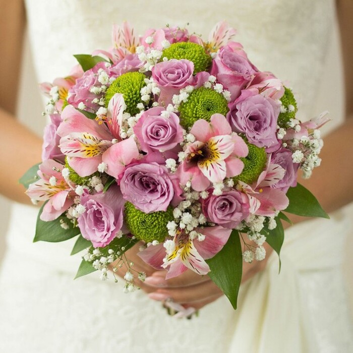 The essence of a South Hackney wedding is captured in this radiant, hand-held bridal bouquet-a full, spherical composition glowing with soft hues and natural vibrancy. Central to the bouquet are roses in gentle shades of blush and lavender, their pristine, velvety petals evoking the tranquil flower beds of Haggerston Park at dawn. Dotted among these are striking lime green button chrysanthemums, providing an energizing, modern element reminiscent of the fresh blooms seen at Broadway Market. Pink alstroemeria lilies appear throughout, their unique trumpet-shaped petals and lively maroon and yellow streaks adding a subtle exotic flair inspired by the area's rich cultural tapestry. Wisps of delicate baby's breath weave seamlessly between the main blooms, lending a whimsical, airy lightness that softens the entire arrangement. A scattering of deep green leaves provides a grounding presence at the base, echoing the lush neighbourhood gardens. The bride's hands-one gently adorned with a classic ring-cradle the stems, wrapped in ribbon, against the flowing white lace of her gown, reflecting the calm joy of a South Hackney celebration. This bouquet, designed by a local florist with a passion for both tradition and innovation, exemplifies the cheerful elegance perfect for modern East London weddings or intimate gatherings by the canals.