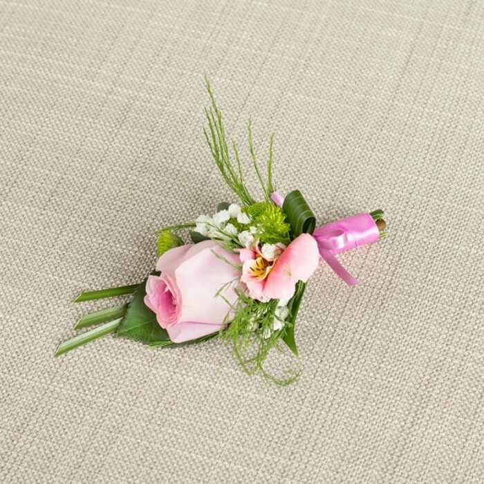 Placed at a gentle angle on a softly woven, light beige linen, this beautifully constructed boutonniere tells a story of South Hackney's creative energy and romantic heart. The principal flower-a tightly wound, blush pink rosebud-anchors the composition, its petals shading from a richer pink core outward to creamy tones at the edge, radiating youth and freshness. Flanking the rose, a subtle Alstroemeria, delicate in peach and pink, reveals its interior banded with faded gold and fragile brown flecks, reminiscent of early spring mornings along Hackney's Regent's Canal. Haloing the arrangement, fine clouds of white baby's breath and bright green button mums are woven with attentiveness, their forms echoing the wilder green spaces throughout Victoria Park. The backing is composed of lively, fern-like sprigs mingled with broader leaves, some folded with deft fingers for architectural effect. Wrapped neatly around the stems, a rich fuchsia pink satin ribbon is tied in a small yet exuberant bow, catching stray morning light in its gleam. Every detail-from the subtle gloss of foliage to the understated neutrality of the textile background-evokes the artistry and precision of local South Hackney florists. This floral pin is designed for life's special occasions, as fitting for a wedding at York Hall as for a refined prom or sophisticated event, suggesting celebration, fresh beginnings, and community artistry.