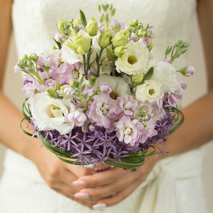 A detailed, close-up image reveals an artfully designed bridal bouquet being gently held by a bride in South Hackney, with her manicured fingers in blush-pink polish delicately cupping the base. Lush white lisianthus flowers with ruffled leaves and creamy, yellow-green centres capture the eye, their elegance emblematic of modern wedding style in Hackney's vibrant community. Clusters of pastel pinks and lilacs-likely fragrant stock-add both colour transition and romantic texture, their frothy layers evoking a sense of softness and joy while recalling the blooms seen at Columbia Road Flower Market. The base of the bouquet is structured by a striking collar of purple star-shaped allium, their geometric presence making a bold, creative statement inspired by local Hackney floristry. Encircling this, deep green bear grass spirals elegantly, providing a refined, architectural contrast to the arrangement's soft upper layers. The partially wrapped stems are supported by the bride's careful hands, hinting at the meticulous attention to detail invested by the area's expert florists. Behind, a soft-focus background reveals a glimpse of a white or ivory wedding dress-perhaps featuring delicate lace-highlighting the bouquet's pride of place in the scene. This bouquet exemplifies South Hackney's artistic floral heritage: a modern masterpiece for weddings and life's most beautiful gatherings, available for local delivery from trusted Hackney florists.