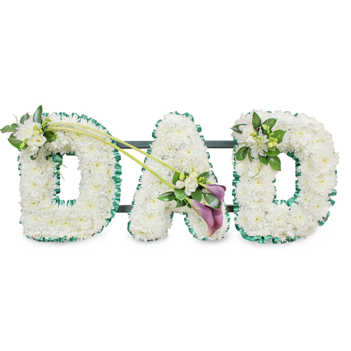 A beautifully composed funeral flower display forms the word "DAD" in bold, standing letters, captured against a bright white background in a style that reflects the quiet, handcrafted work of local florists in South Hackney. Each letter is sculpted from a bed of tight white chrysanthemums, the compact blooms arranged so closely that they create a uniform, cushion-like texture. The petals are crisp and flawless, their soft curves suggesting purity and peace, as if offering a gentle place for memories to rest. Around the outside edge of each three-dimensional letter, a deep green ribbon is carefully folded into ruffles, tracing every angle and rounded section with a precise, satin sheen. This rich green border frames the white flowers beautifully, evoking the calm of nearby green spaces like Victoria Park and the tree-lined paths that weave through the neighbourhood. Behind the letters, discrete horizontal dark bars of the support frame are visible, indicating that the arrangement is designed to stand upright through a service or at a graveside. Elegant finishing touches add character: a modest cluster of white freesia and fresh foliage is nestled on the upper left side of the first "D", while a similar, slightly asymmetric spray rests on the upper right side of the last "D". Between these, from the top of the first "D" down to the lower right of the central "A", two long, arching green stems sweep diagonally, leading the eye to a focal spray at the base of the "A". Here, two deep purple calla lilies, velvety and trumpet-shaped, sit amid more white freesia blossoms and glossy green leaves. The interplay of the dark, dignified purple with the tranquil white and green tones creates a tribute that feels both traditional and deeply emotional - a loving, respectful farewell to a father figure, suited to funerals, memorials, or remembrance gatherings across South Hackney.