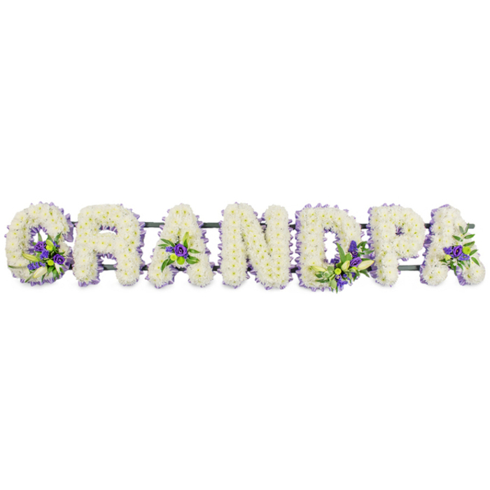 In this photograph, the word "GRANDPA" is lovingly spelled out in large floral letters, laid side by side in a horizontal line that would look at home in a South Hackney chapel or crematorium. Each letter is constructed on a hidden base and completely blanketed with small white funeral blooms-most likely chrysanthemums or carnations-placed so closely that they form a solid, cushion-like mass of petals. The flowers have a soft, slightly ruffled texture, and their uniform whiteness creates a quiet, almost luminous surface that suggests peace and remembrance. Within this field of white, the florist has introduced pockets of colour using deep purple and soft lavender flowers, such as lisianthus or petite chrysanthemums, accompanied by fresh green foliage. These accents are arranged thoughtfully across the tribute: gathered at the base of the "G," tucked into the open centres of each "A," enhancing the vertical and curved parts of the "P," and appearing subtly in other letters to maintain visual balance. The interplay between crisp white, regal purple, and vibrant green brings gentle movement to the arrangement without disturbing its dignified calm. Beneath and behind the letters lies a narrow, dark green support bar that joins the individual forms into one continuous word, keeping everything aligned and ready for display on a coffin or stand during the service. The overall mood is respectful and intimate, the kind of custom tribute families in South Hackney might choose when they want to see "Grandpa" written clearly in flowers-perhaps in memory of a man who enjoyed quiet walks around Victoria Park or watching the world go by along Mare Street. It offers a beautiful, tangible way to say farewell, wrapped in softness and care.
