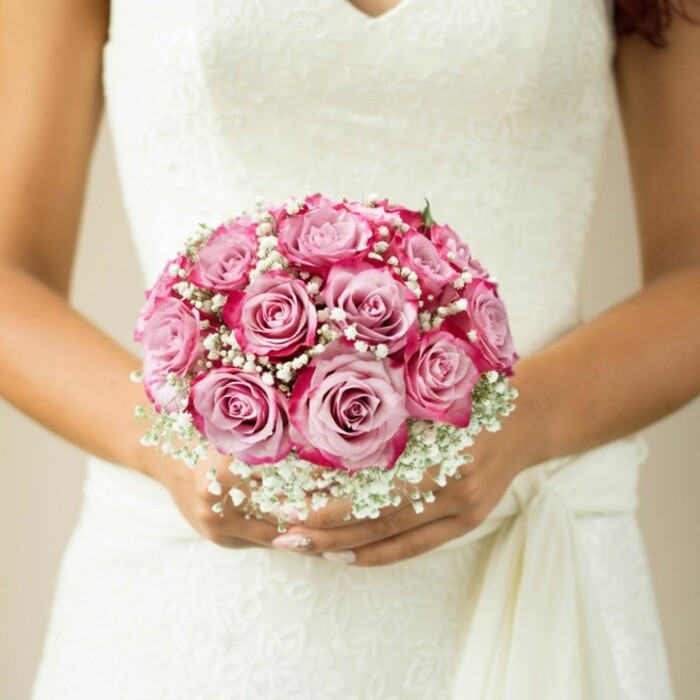 The image captures an intimate South Hackney wedding moment, focusing closely on a bride's hands as they cradle a lush, meticulously arranged bridal bouquet. The bouquet, hand-tied into a perfect round, is made up of medium-sized roses whose petals open in layers from soft pink at the centres to a deep, electric magenta flowing to their outermost tips. Interspersed through the roses are delicate sprigs of baby's breath-cloudlike, white, and airy-providing gentle texture and classical charm. Near the bouquet's top, a hint of subtle greenery peeks out, a nod to the natural beauty found along Regent's Canal. The bride's hands-appearing sun-kissed and wearing a polished pale-pink manicure-support the blooms just above the sash of her gown. Her white wedding dress, detailed with intricate floral lace and a softly plunging V-neck, is gathered at one side, the drape of fabric conjuring a sense of movement and grace. Against a muted, blurred background, the bouquet radiates warmth, local artistry, and a feeling of romantic anticipation-an ideal vision for weddings or bridal inspiration in South Hackney.